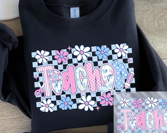 May include: Black sweatshirt with a colorful floral and checkerboard design. The word "Teacher" is in a bubble letter style with pink, blue, and white accents. A pair of scissors and a pencil are also included in the design.