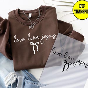 May include: A brown sweatshirt with the phrase "love like jesus" in white script and a bow graphic. A silver necklace with two charms is visible. A DTF transfer is next to the sweatshirt. The sweatshirt is folded on top of blue jeans.