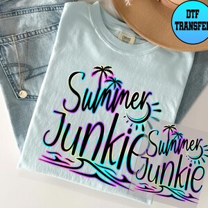 Puede incluir: Una camiseta azul claro con un gr&aacute;fico negro que dice "Summer Junkie" en un degradado arco&iacute;ris. El gr&aacute;fico tambi&eacute;n incluye palmeras, olas y un sol. La camiseta est&aacute; sobre una mesa con un par de pantalones vaqueros azules y un sombrero marr&oacute;n.