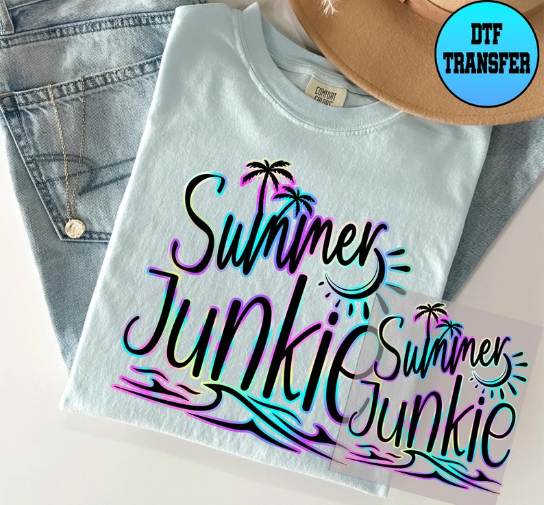 Puede incluir: Una camiseta azul claro con un gr&aacute;fico negro que dice "Summer Junkie" en un degradado arco&iacute;ris. El gr&aacute;fico tambi&eacute;n incluye palmeras, olas y un sol. La camiseta est&aacute; sobre una mesa con un par de pantalones vaqueros azules y un sombrero marr&oacute;n.