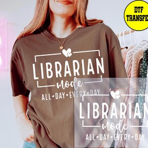 May include: A brown t-shirt with the text "LIBRARIAN mode ALL DAY EVERY DAY" in white. The design includes a heart and a rectangular frame. A yellow circle with "DTF TRANSFER" is in the upper right corner.