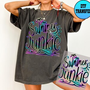 Puede incluir: Una camiseta gris oscura con un dise&ntilde;o degradado arco&iacute;ris que dice "Summer Junkie" con palmeras y un gr&aacute;fico de sol. El dise&ntilde;o est&aacute; en una hoja de transferencia transparente.