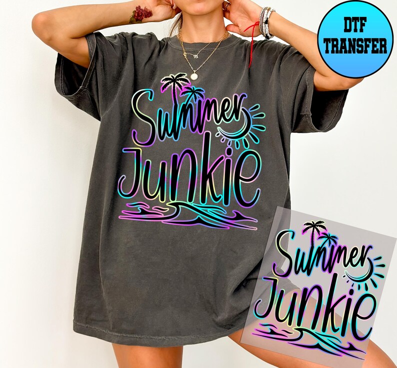 Puede incluir: Una camiseta gris oscura con un dise&ntilde;o degradado arco&iacute;ris que dice "Summer Junkie" con palmeras y un gr&aacute;fico de sol. El dise&ntilde;o est&aacute; en una hoja de transferencia transparente.