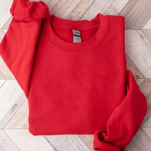 May include: A bright red crewneck sweatshirt, folded neatly. The sweatshirt is made of a soft material and has a classic, casual style. The sleeves are folded, and the Gildan tag is visible.