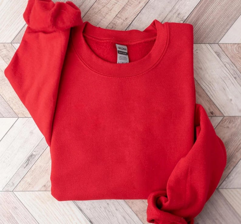 May include: A bright red crewneck sweatshirt, folded neatly. The sweatshirt is made of a soft material and has a classic, casual style. The sleeves are folded, and the Gildan tag is visible.