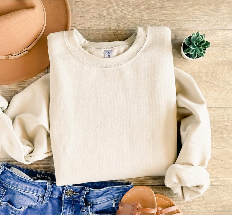 May include: A cream-colored crewneck sweatshirt with rolled-up sleeves, paired with blue denim jeans, brown sandals, and a tan hat. A small potted succulent is also visible. The items are arranged on a light wood-grain surface.