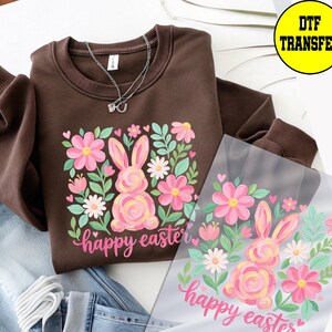 May include: A brown sweatshirt with a floral and bunny design. The design includes pink flowers, green leaves, and a pink bunny with the words "happy easter" in pink script. A silver necklace is on the sweatshirt.