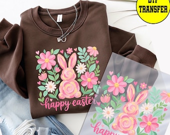 May include: A brown sweatshirt with a floral and bunny design. The design includes pink flowers, green leaves, and a pink bunny with the words "happy easter" in pink script. A silver necklace is on the sweatshirt.