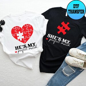May include: Two t-shirts, one white and one black, with matching designs. The white shirt features a red heart made of puzzle pieces with the text "HE'S MY perfect match". The black shirt has a red puzzle piece with the text "SHE'S MY perfect match".