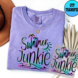 Puede incluir: Una camiseta morada con el texto "Summer Junkie" en negro con un contorno de arco&iacute;ris. El texto est&aacute; sobre un fondo blanco con una palmera, un sol y olas. La camiseta est&aacute; sobre una superficie blanca con un sombrero marr&oacute;n y una pieza de follaje seco.