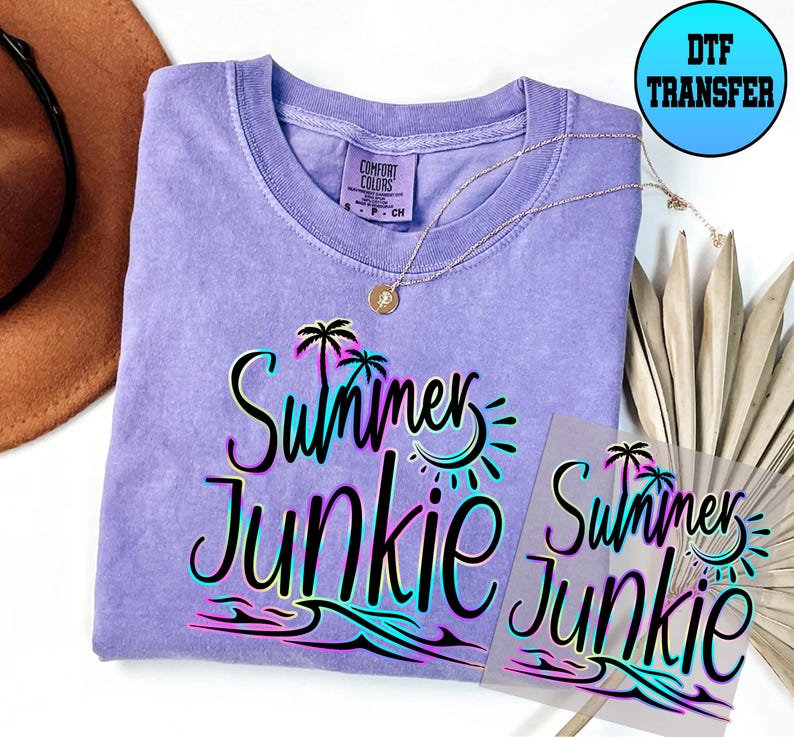 Puede incluir: Una camiseta morada con el texto "Summer Junkie" en negro con un contorno de arco&iacute;ris. El texto est&aacute; sobre un fondo blanco con una palmera, un sol y olas. La camiseta est&aacute; sobre una superficie blanca con un sombrero marr&oacute;n y una pieza de follaje seco.