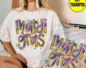May include: Cream-colored t-shirt with a colorful "Mardi Gras" design. The text is in purple, gold, and green, with decorative accents. A DTF transfer is visible in the image.