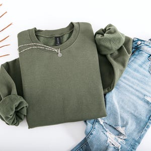 May include: An olive green sweatshirt with a silver necklace featuring a circular pendant. The sweatshirt is paired with light blue ripped jeans. The image is shot from above on a white surface, showcasing a casual outfit.