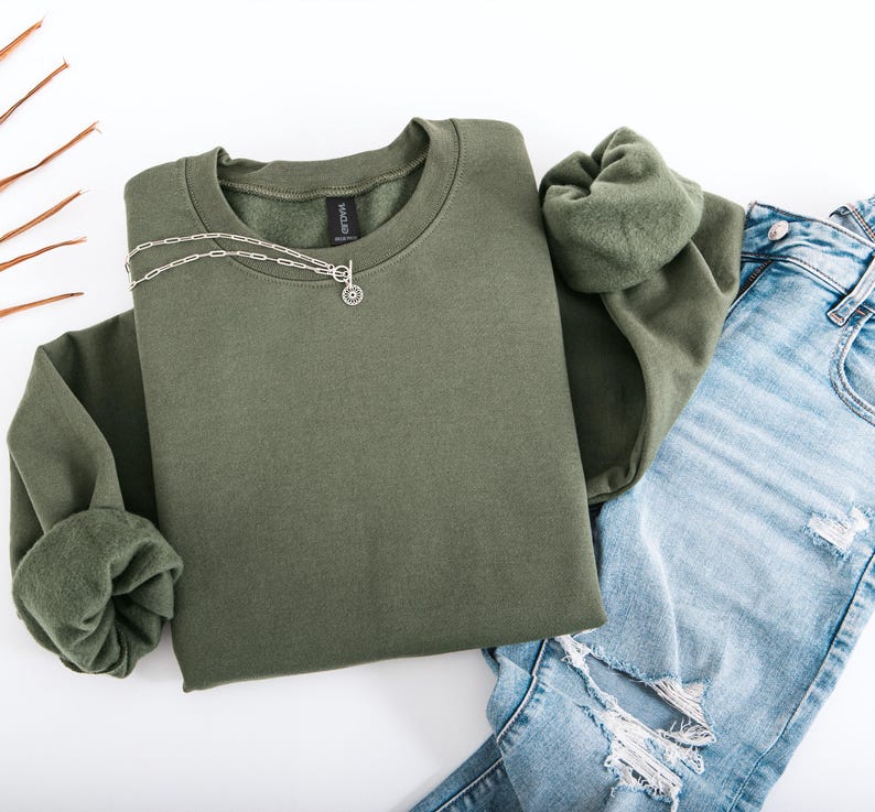 May include: An olive green sweatshirt with a silver necklace featuring a circular pendant. The sweatshirt is paired with light blue ripped jeans. The image is shot from above on a white surface, showcasing a casual outfit.