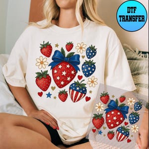 May include: A white t-shirt with a red, white, and blue strawberry design. The strawberries are decorated with stars and have a patriotic theme. The shirt is perfect for summer and the 4th of July.