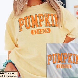 K&ouml;nnte beinhalten: Ein hellgelbes Kurzarm-T-Shirt mit dem Text PUMPKIN SEASON in einer abgenutzten orangefarbenen Schrift mit schwarzer Umrandung. Das Design ist in einem gro&szlig;en Bogen auf der Brust gedruckt. Eine separate quadratische Grafik zeigt das Motiv.