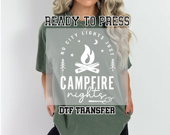 May include: Green t-shirt with a white graphic design that says "No city lights just campfire nights" with a campfire and trees. The text "DTG Transfer" is also on the shirt.