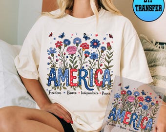 May include: Cream-colored t-shirt featuring a floral design with the word "AMERICA" in red and blue. The design includes flowers, butterflies, and the words "Freedom, Brave, Independence, Power."
