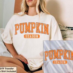 K&ouml;nnte beinhalten: Ein cremefarbenes Kurzarm-T-Shirt mit dem orangefarbenen, abgenutzten Schriftzug PUMPKIN SEASON in College-Schriftart. Das Design zeigt das Wort PUMPKIN in einem gro&szlig;en Bogen &uuml;ber dem Wort SEASON in einem orangefarbenen rechteckigen Rahmen.