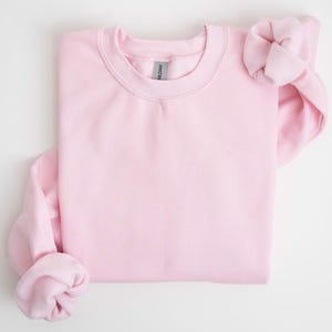 May include: A light pink crewneck sweatshirt, folded neatly. The sleeves are cuffed, and the fabric appears soft. The Gildan brand tag is visible at the neckline. The sweatshirt is a solid color, suitable for casual wear.