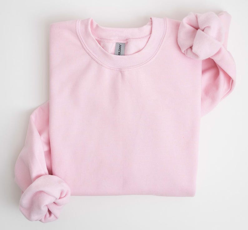 May include: A light pink crewneck sweatshirt, folded neatly. The sleeves are cuffed, and the fabric appears soft. The Gildan brand tag is visible at the neckline. The sweatshirt is a solid color, suitable for casual wear.