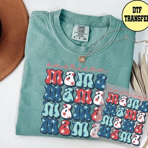 May include: A teal Comfort Colors t-shirt with the words "American Mama" in red, white, and blue bubble letters. A silver necklace with a pendant rests on the shirt. A DTF transfer is visible.