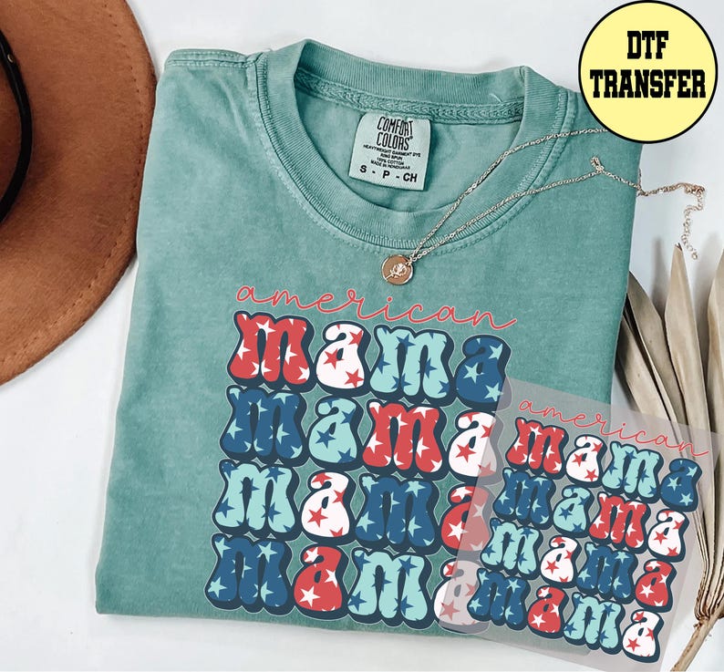 May include: A teal Comfort Colors t-shirt with the words "American Mama" in red, white, and blue bubble letters. A silver necklace with a pendant rests on the shirt. A DTF transfer is visible.