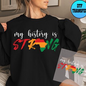 May include: Black sweatshirt with the text "my history is STRONG" in white with a red, yellow, and green outline of the continent of Africa. The sweatshirt is being modelled by a person.