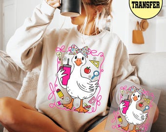 May include: Cream-colored sweatshirt featuring a cartoon goose with a floral bow, holding a pink tumbler. The goose is surrounded by school-themed graphics like a pencil, apple, and ruler. A DTF TRANSFER logo is visible.