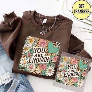 May include: A brown sweatshirt with a floral graphic that reads "YOU ARE ENOUGH." The design includes a green heart, pink roses, and daisies. A DTF transfer is also visible.