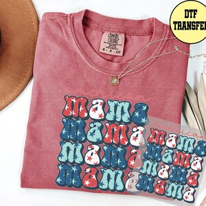 May include: A red t-shirt with the words "american mama" in a retro font, with red, white, and blue stars. A DTF transfer is visible. A gold necklace is draped on the shirt.