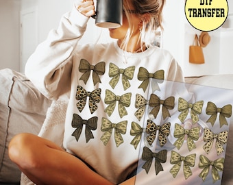 May include: A cream-colored sweatshirt featuring a repeating pattern of various bow designs. The bows are in shades of brown, olive green, and leopard print. The text "DTF TRANSFER" is in a yellow circle in the upper right corner.