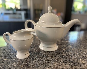 May include: A white porcelain tea set with a teapot and creamer. The teapot has a lid and spout, and the creamer has a handle. Both pieces are decorated with a silver trim.