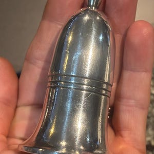 Könnte beinhalten: Ein kleiner, silberfarbener, glockenförmiger Gegenstand mit abgerundeter Oberseite und ausgestelltem Boden. Der Gegenstand hat eine glatte, reflektierende Oberfläche und scheint aus Metall zu sein. Die Oberseite hat einen kleinen, dekorativen Knopf. Die Glocke wird in der Hand einer Person gehalten.