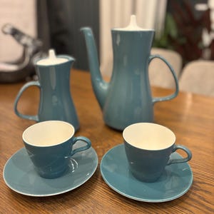 Puede incluir: Un juego de té de cerámica azul verdoso con una tetera, una jarra de crema y dos tazas y platillos.