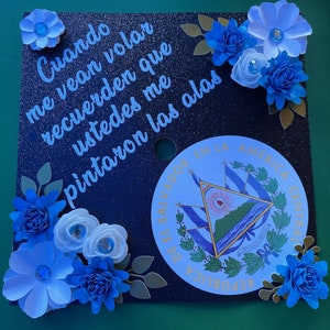 Peut inclure: Un bonnet de graduation noir décoré de fleurs en papier bleu et blanc. Le bonnet porte une inscription blanche qui dit "Cuando me vean volar recuerden que ustedes me pintaron las alas". Le bonnet comporte également un autocollant circulaire avec les armoiries d'El Salvador.