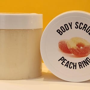Op de afbeelding: Een witte pot met een wit deksel met een lichaamsscrub. Het deksel heeft een perzikring snoepje erop gedrukt met de tekst "BODY SCRUB PEACH RING".
