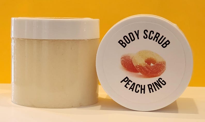 Op de afbeelding: Een witte pot met een wit deksel met een lichaamsscrub. Het deksel heeft een perzikring snoepje erop gedrukt met de tekst "BODY SCRUB PEACH RING".