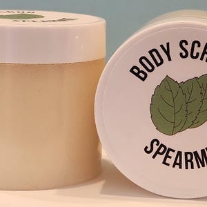 Op de afbeelding: Een witte pot met een wit deksel gevuld met een lichtbruine bodyscrub. Het deksel heeft een zwart label met de tekst "BODY SCRUB SPEARMINT" met een groene illustratie van een muntblad.