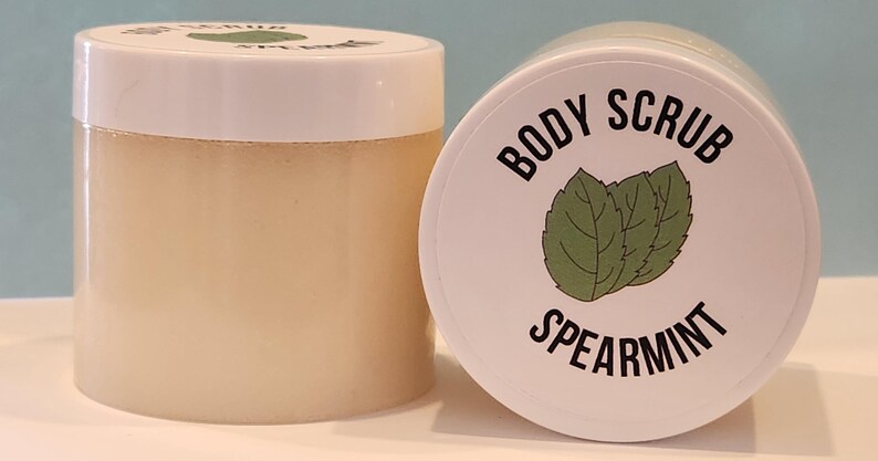 Op de afbeelding: Een witte pot met een wit deksel gevuld met een lichtbruine bodyscrub. Het deksel heeft een zwart label met de tekst "BODY SCRUB SPEARMINT" met een groene illustratie van een muntblad.