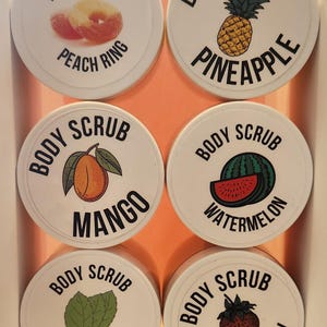 Op de afbeelding: Zes witte ronde containers met zwarte deksels en labels. Elke container heeft een ander fruitontwerp en de tekst "Body Scrub" met de naam van het fruit. De vruchten zijn perzikring, ananas, mango, watermeloen, munt en aardbei.
