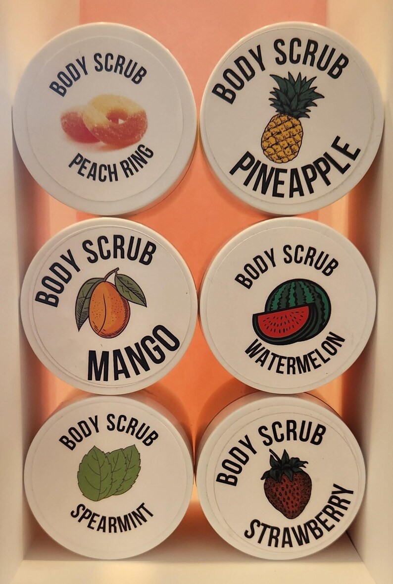 Op de afbeelding: Zes witte ronde containers met zwarte deksels en labels. Elke container heeft een ander fruitontwerp en de tekst "Body Scrub" met de naam van het fruit. De vruchten zijn perzikring, ananas, mango, watermeloen, munt en aardbei.