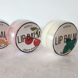 May include: Three small round lip balm containers with white labels and different fruit and herb designs. The labels read "Lip Balm" with the flavor listed below. The flavors are "Citrus Dream", "Cherry Dream", and "Spearmint".