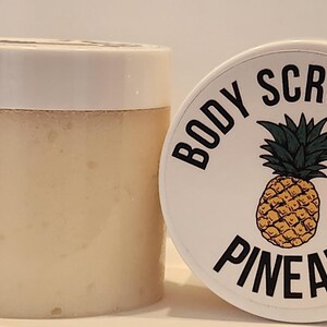 Op de afbeelding: Een witte pot met een zwart-wit etiket waarop "BODY SCRUB PINEAPPLE" staat. Het etiket heeft een tekening van een ananas.