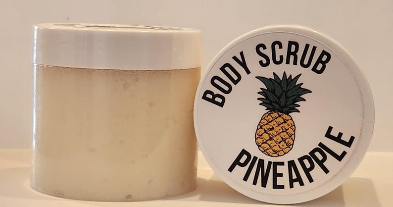 Op de afbeelding: Een witte pot met een zwart-wit etiket waarop "BODY SCRUB PINEAPPLE" staat. Het etiket heeft een tekening van een ananas.