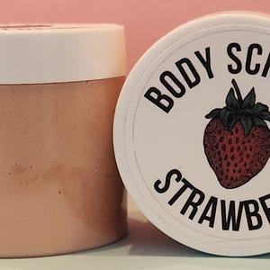 Op de afbeelding: Een witte pot met een roze deksel gevuld met een aardbeien bodyscrub. De pot heeft een wit etiket met de tekst "BODY SCRUB STRAWBERRY" en een rode aardbei-illustratie.