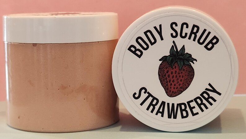 Op de afbeelding: Een witte pot met een roze deksel gevuld met een aardbeien bodyscrub. De pot heeft een wit etiket met de tekst "BODY SCRUB STRAWBERRY" en een rode aardbei-illustratie.