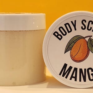 Op de afbeelding: Een witte pot met een wit deksel met een lichtbeige body scrub. Het deksel heeft een zwart-wit label met de tekst "Body Scrub Mango". Het label heeft een cartoonillustratie van een mango.