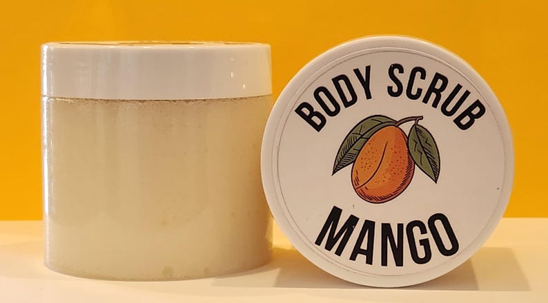 Op de afbeelding: Een witte pot met een wit deksel met een lichtbeige body scrub. Het deksel heeft een zwart-wit label met de tekst "Body Scrub Mango". Het label heeft een cartoonillustratie van een mango.