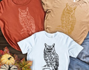 May include: Three t-shirts with different colored backgrounds featuring a black and white illustration of an owl. The owl is facing forward with its wings folded. The t-shirts are laid out on a wooden surface with a pair of blue jeans and pumpkins in the background.